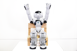 ロボット「NAO」が椅子に座って両腕を真上に上げる体操をしている写真