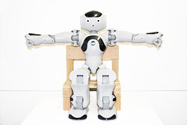 ロボット「NAO」が椅子に座って両腕を左右に上げる体操をしている写真